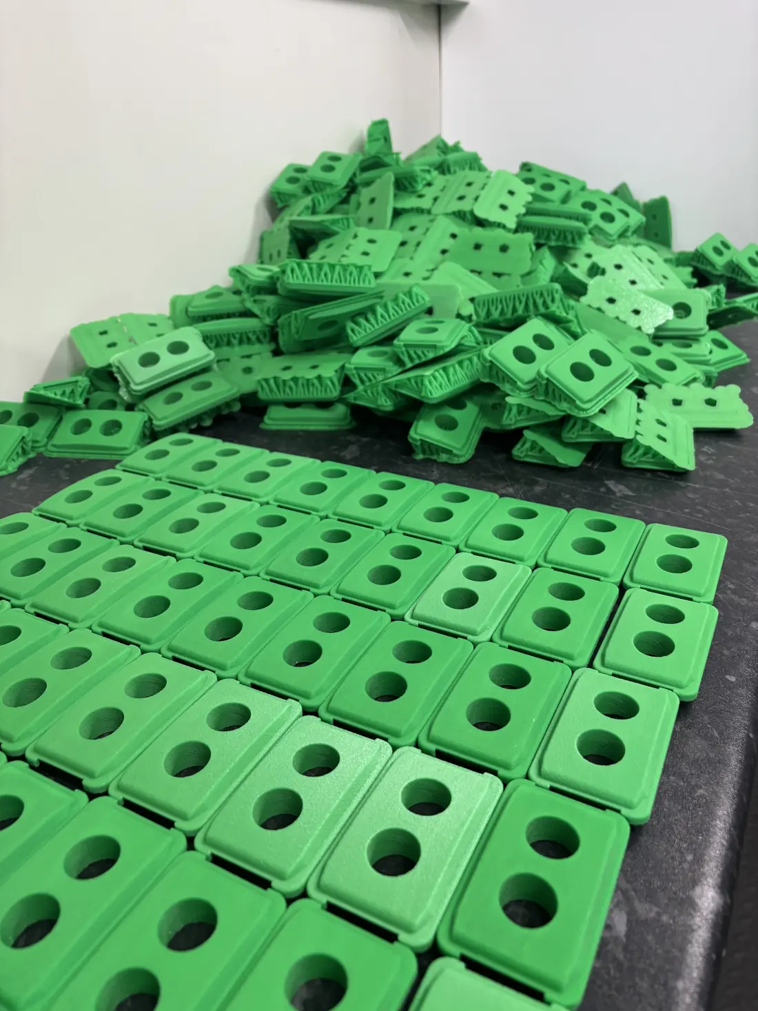 A production run of green 3D printed parts at 3D Printing Express, finished units laid out in rows in front of a larger pile still to be sorted