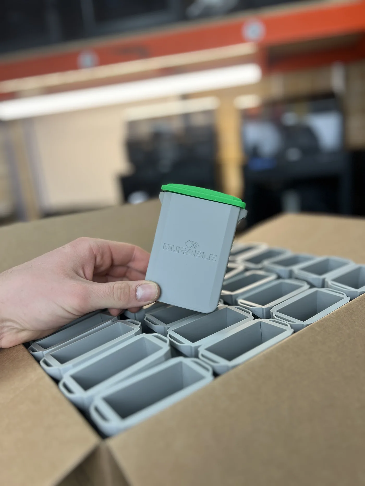 Hand holding a 3D-printed Durable-branded grey pen holder with green lid, over a box filled with the rest of a 500-unit batch run, in the 3D Printing Express workshop