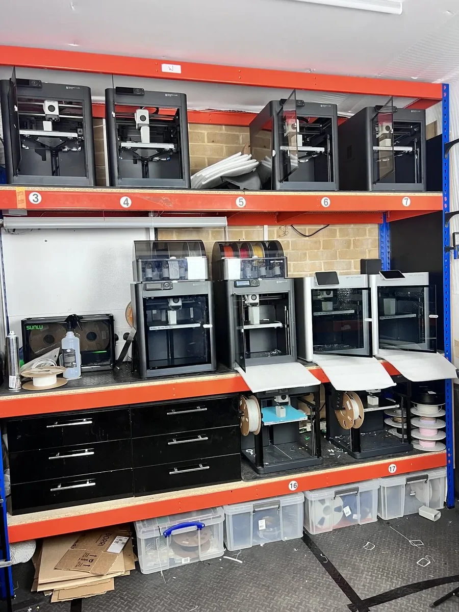 Numbered printer bays on racking in the 3D Printing Express workshop, each bay holding a machine, with filament and parts bins stored around them