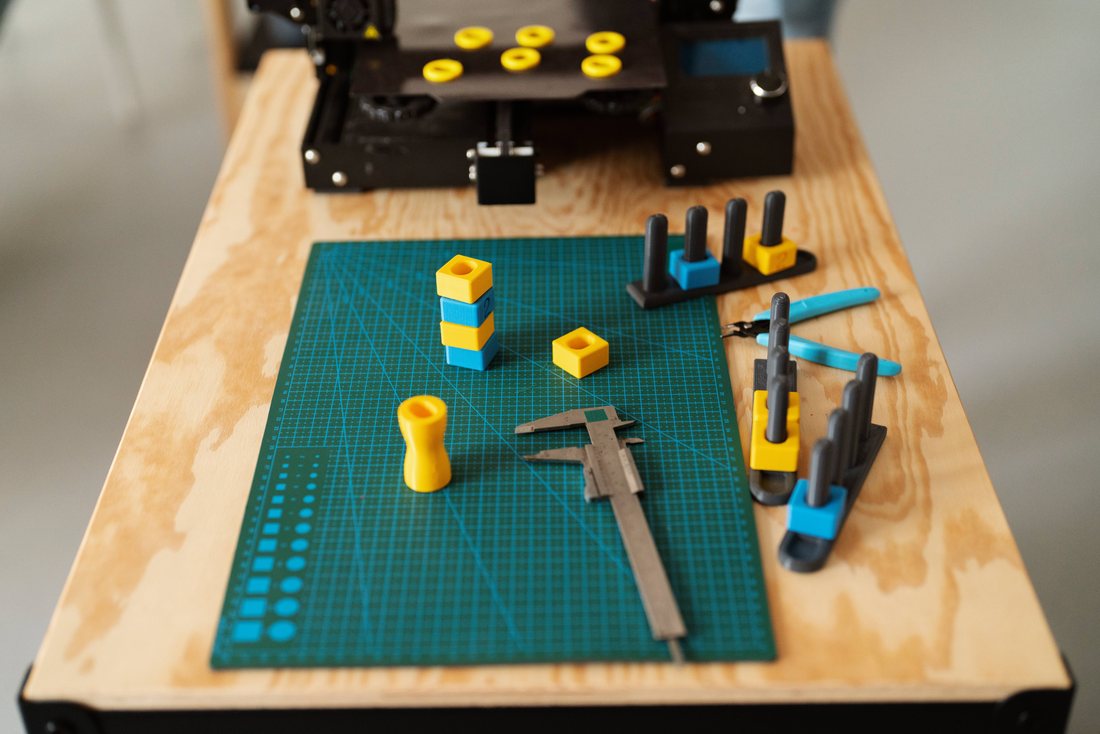 3DPE product-developer bench · real digital calipers and 3D-printed test parts on a cutting mat, with a 3D printer running in the background