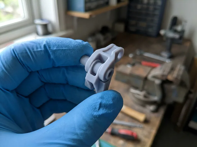 Illustration: a small pale grey resin printed component held in gloved fingertips, showing crisp fine detail and a smooth surface with no visible layer lines