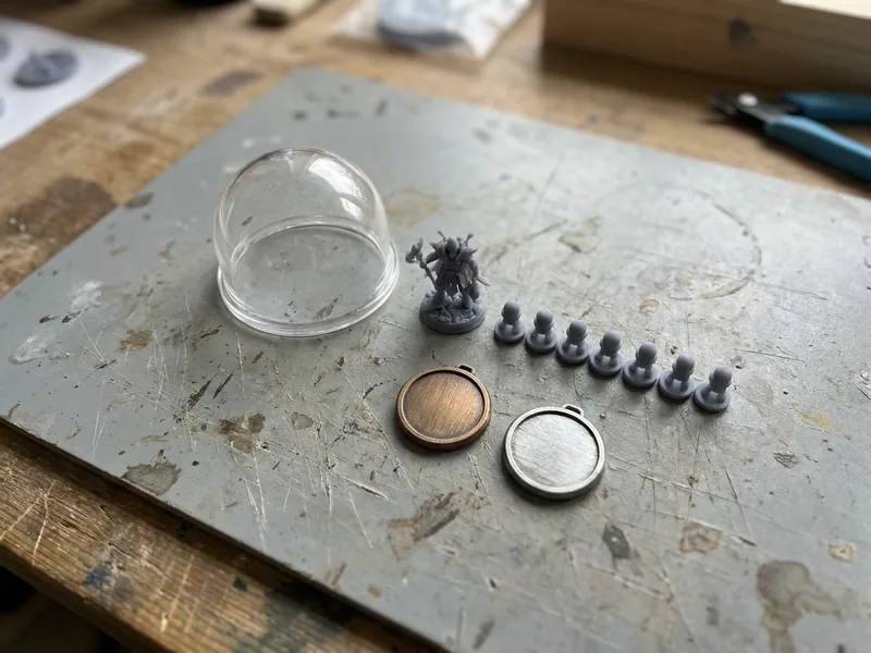 Illustration: a group of small resin printed parts on a bench, a clear cover, a figure part and a set of game pieces